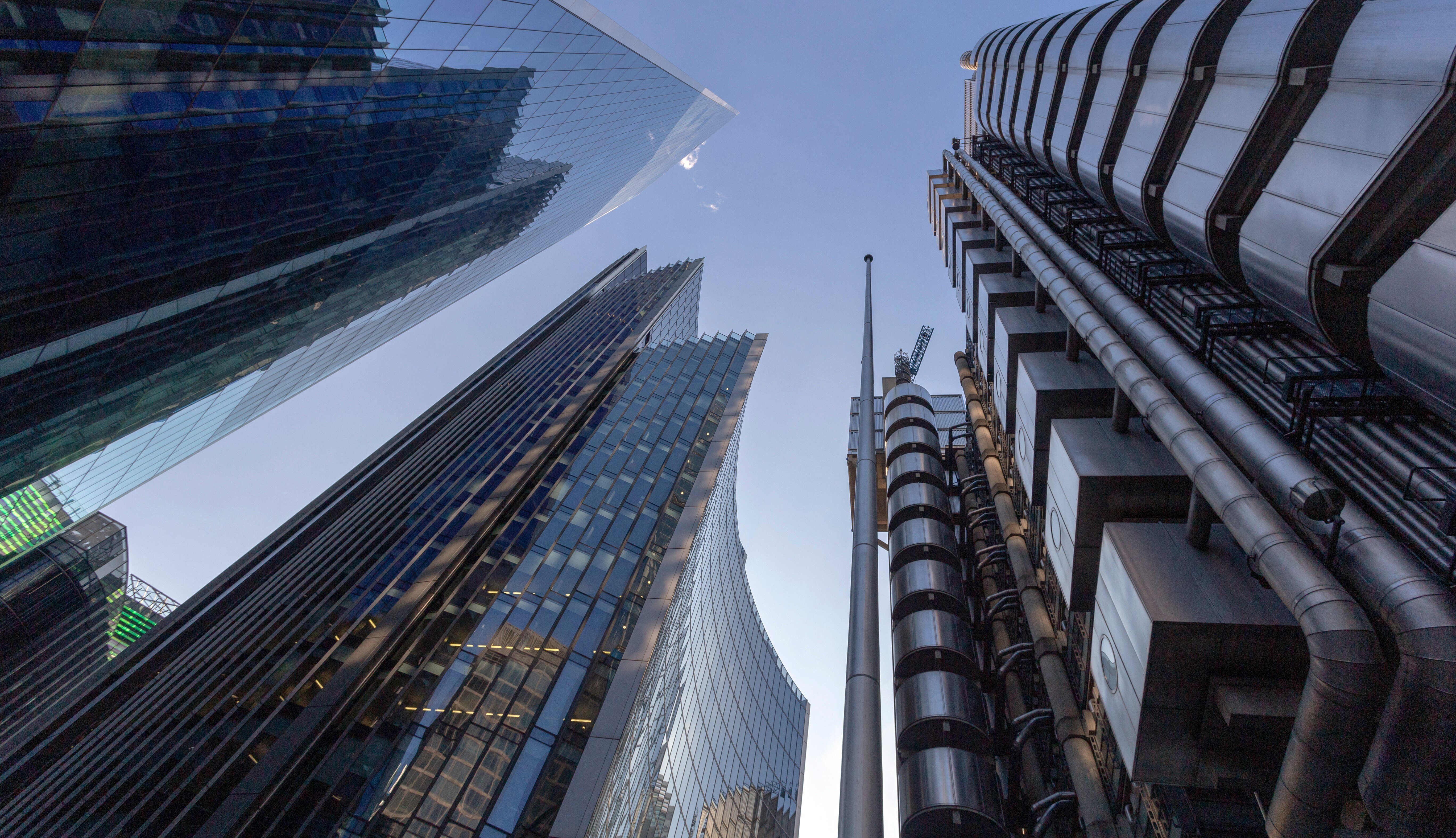 Upward view of modern skyscrapers and architectural buildings against a clear blue sky, showcasing a blend of reflective glass surfaces and industrial steel structures.