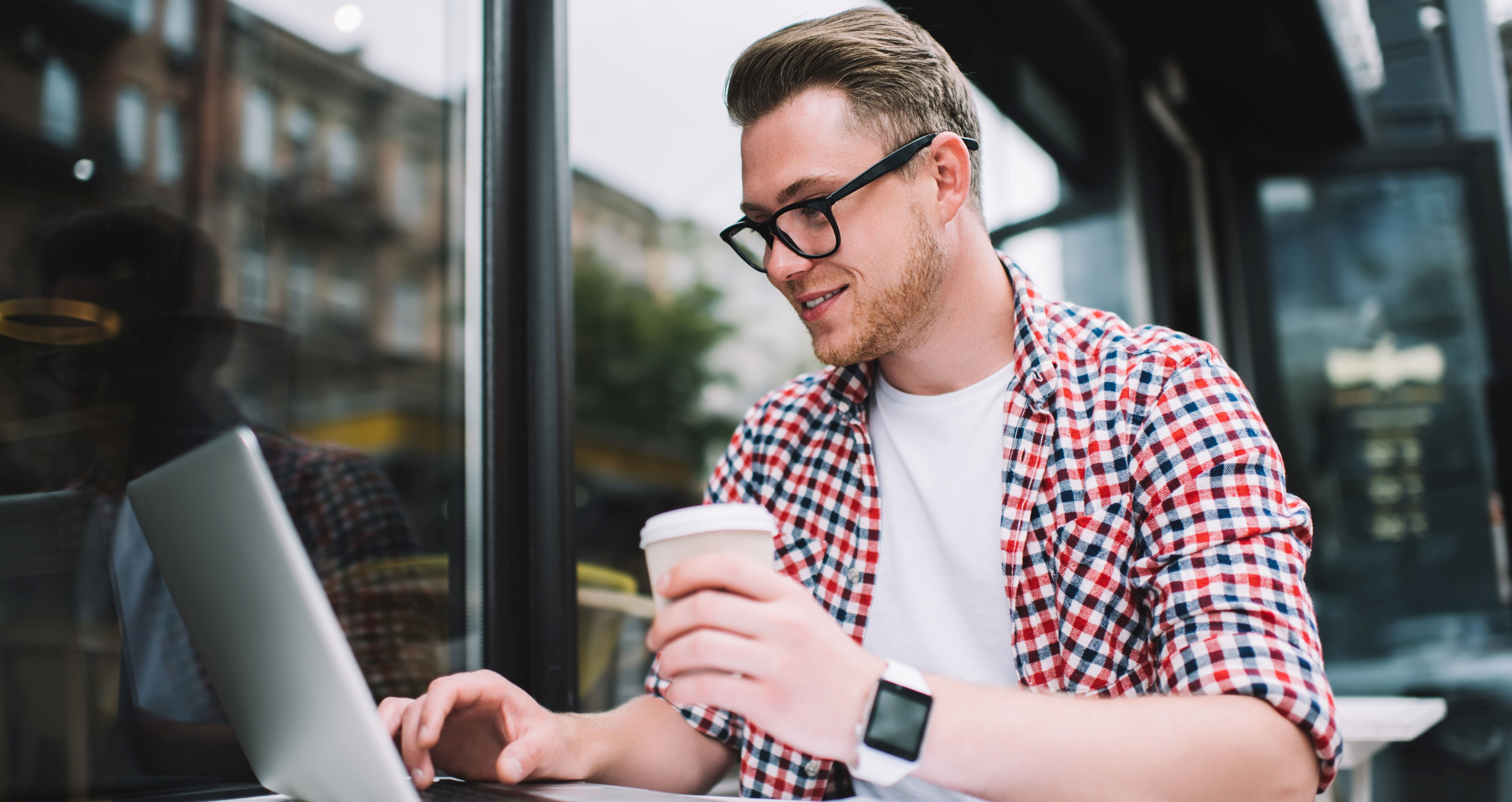 Young man wearing glasses and a plaid shirt, sitting at an outdoor cafe table with a laptop. He is holding a takeaway coffee cup in one hand and smiling at the screen. A notepad and pen are on the table, and he appears to be working or studying.