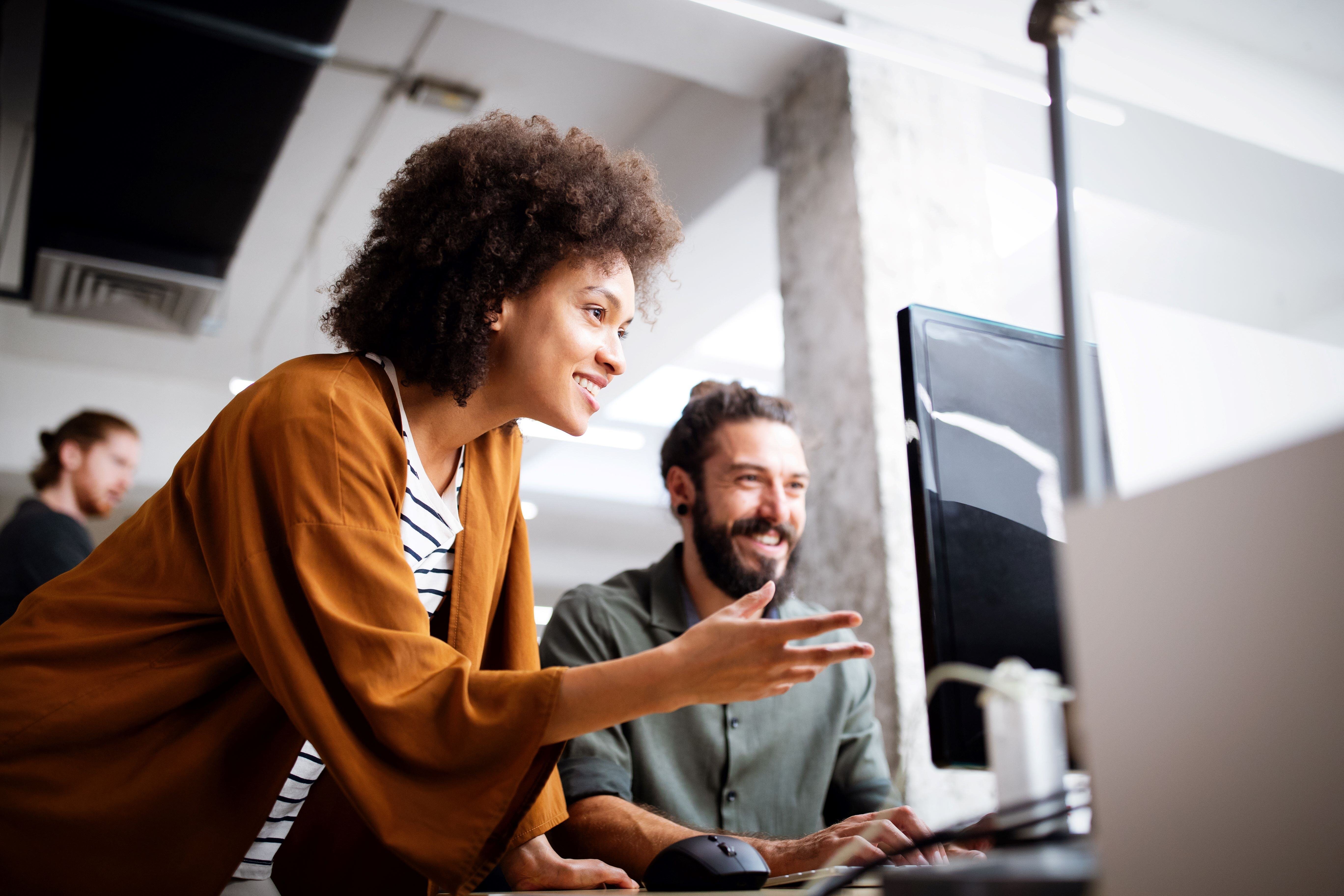 Two colleagues smiling and collaborating at a computer in a bright, modern office. One person is pointing at the screen while the other is using the mouse, indicating a productive and positive work environment.