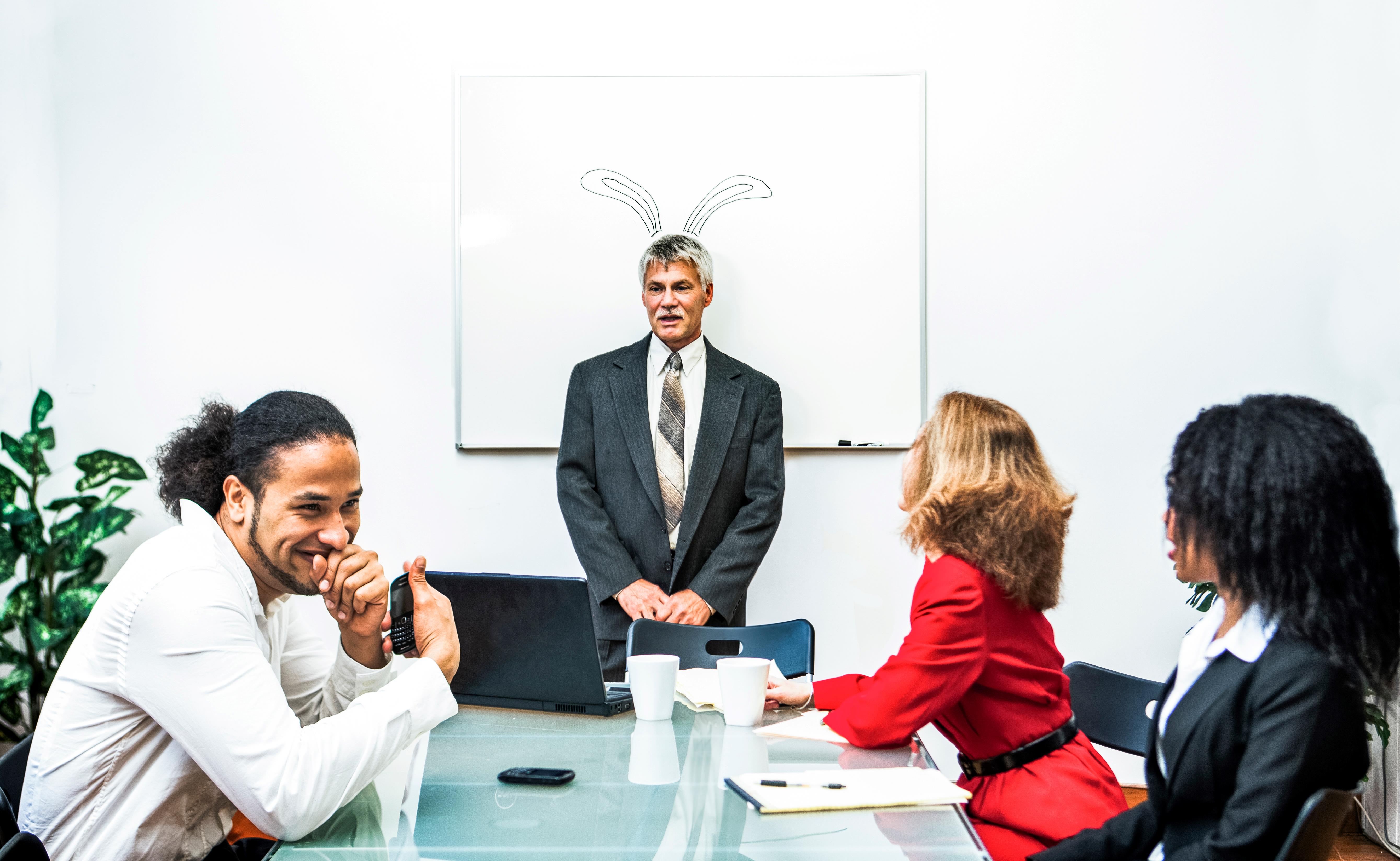 Office workers laughing whilst team leader stands in front of white board with bunny ears behind him